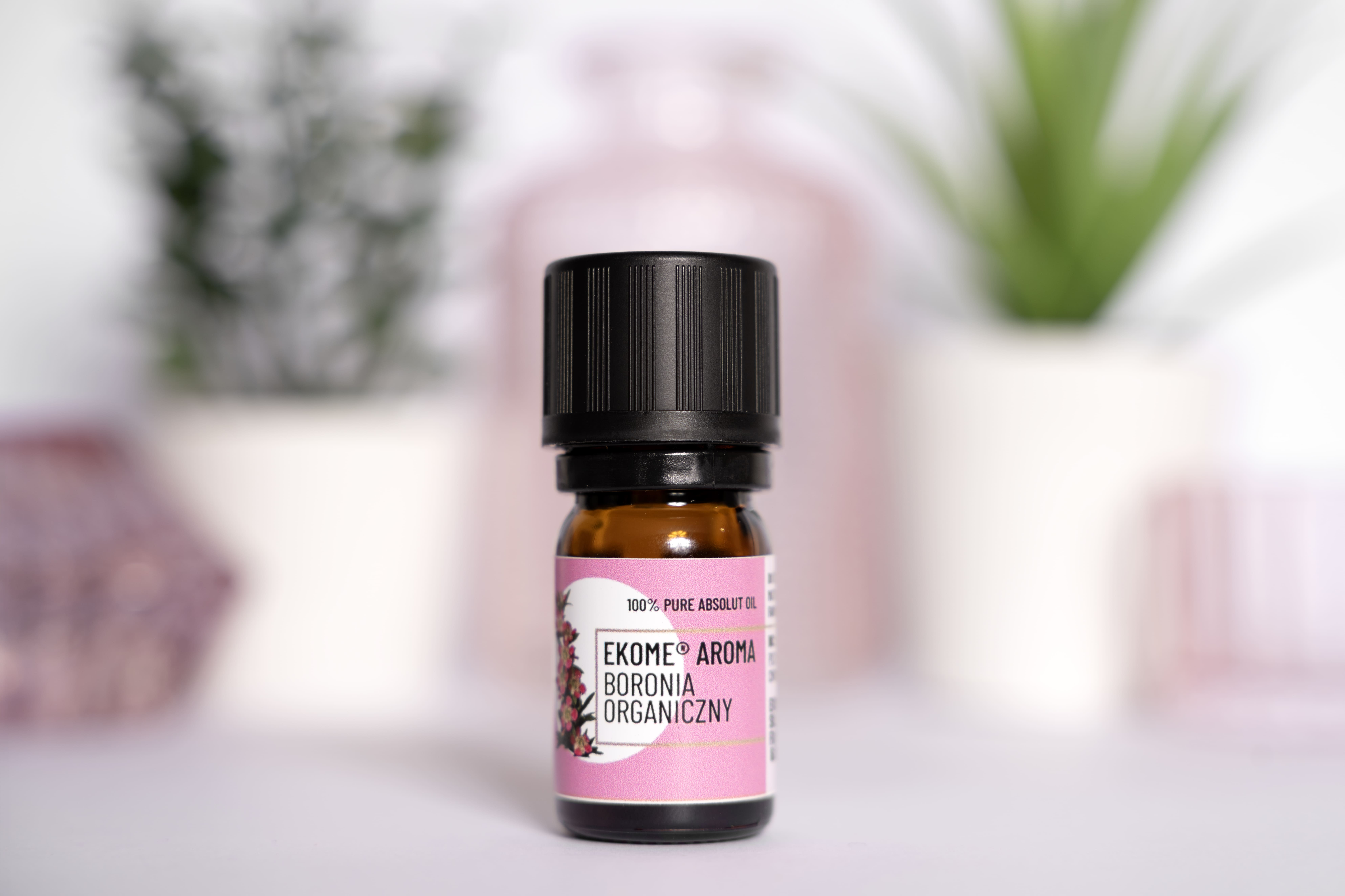 Ekome® Aroma Boronia Organiczny, 100% olejek absolutny, w szklanej buteleczce, ustawiony na jasnym tle z rozmytym tłem roślinnym w tle.