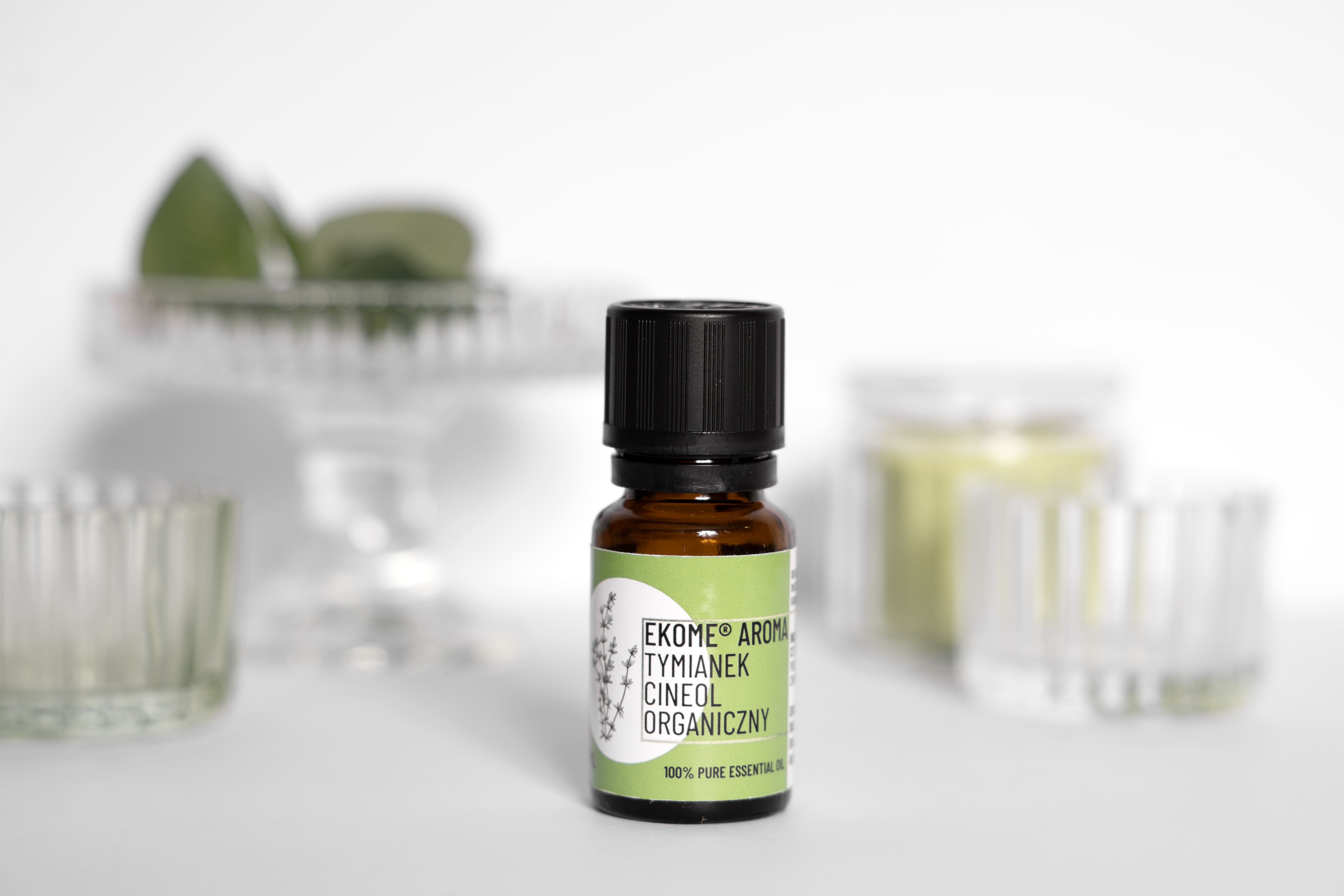 Butelka olejku Ekome Aroma Tymianek Cineol Organiczny na jasnym podłożu, otoczona szklanymi naczyniami.