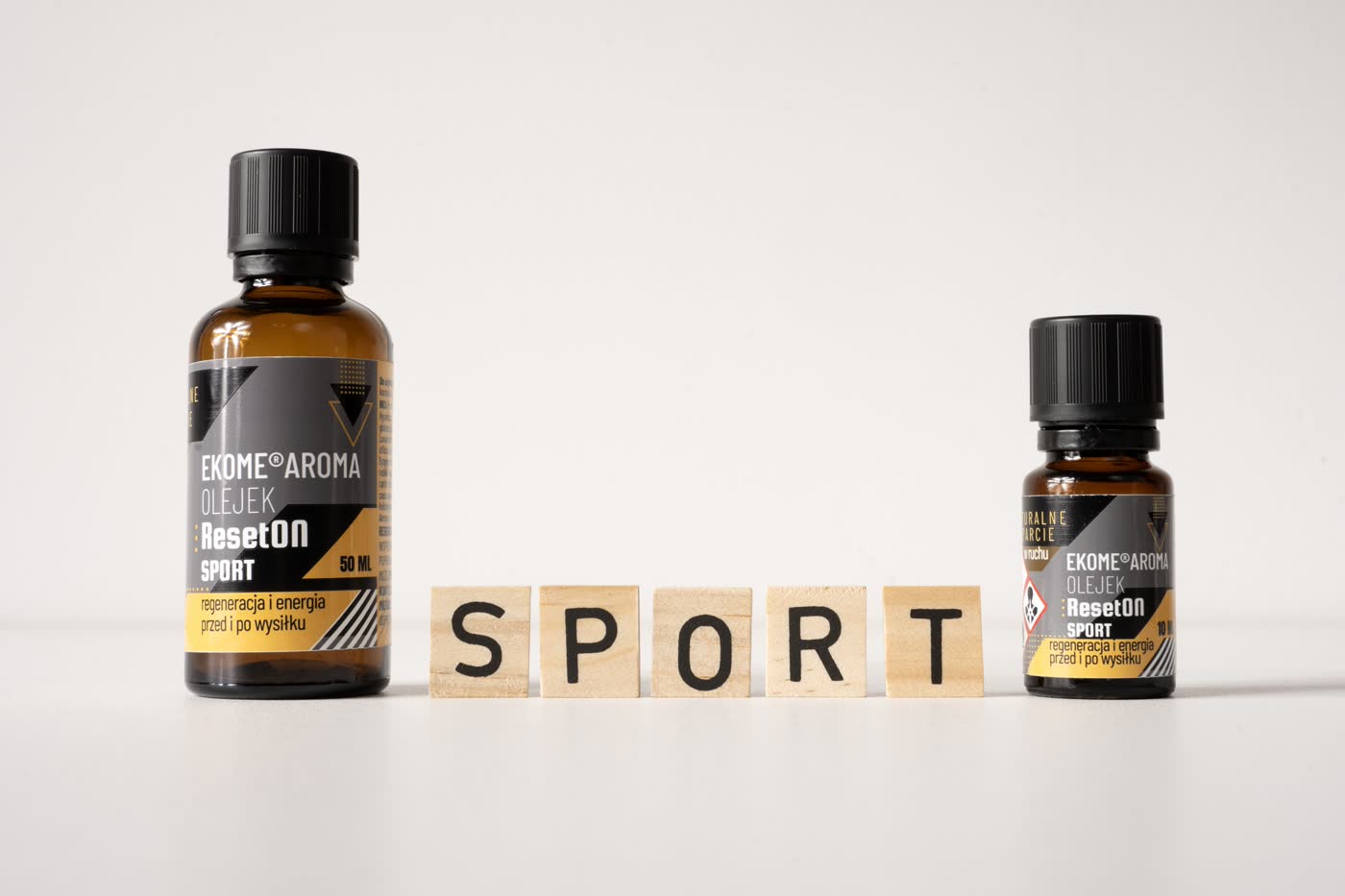 Butelki olejku aromaterapeutycznego Ekome® ResetON Sport w dwóch pojemnościach – 50 ml i 10 ml – ustawione na białym tle, obok drewnianych kostek układających się w napis „SPORT”; etykiety podkreślają działanie regenerujące i wspierające przed oraz po wysiłku fizycznym.