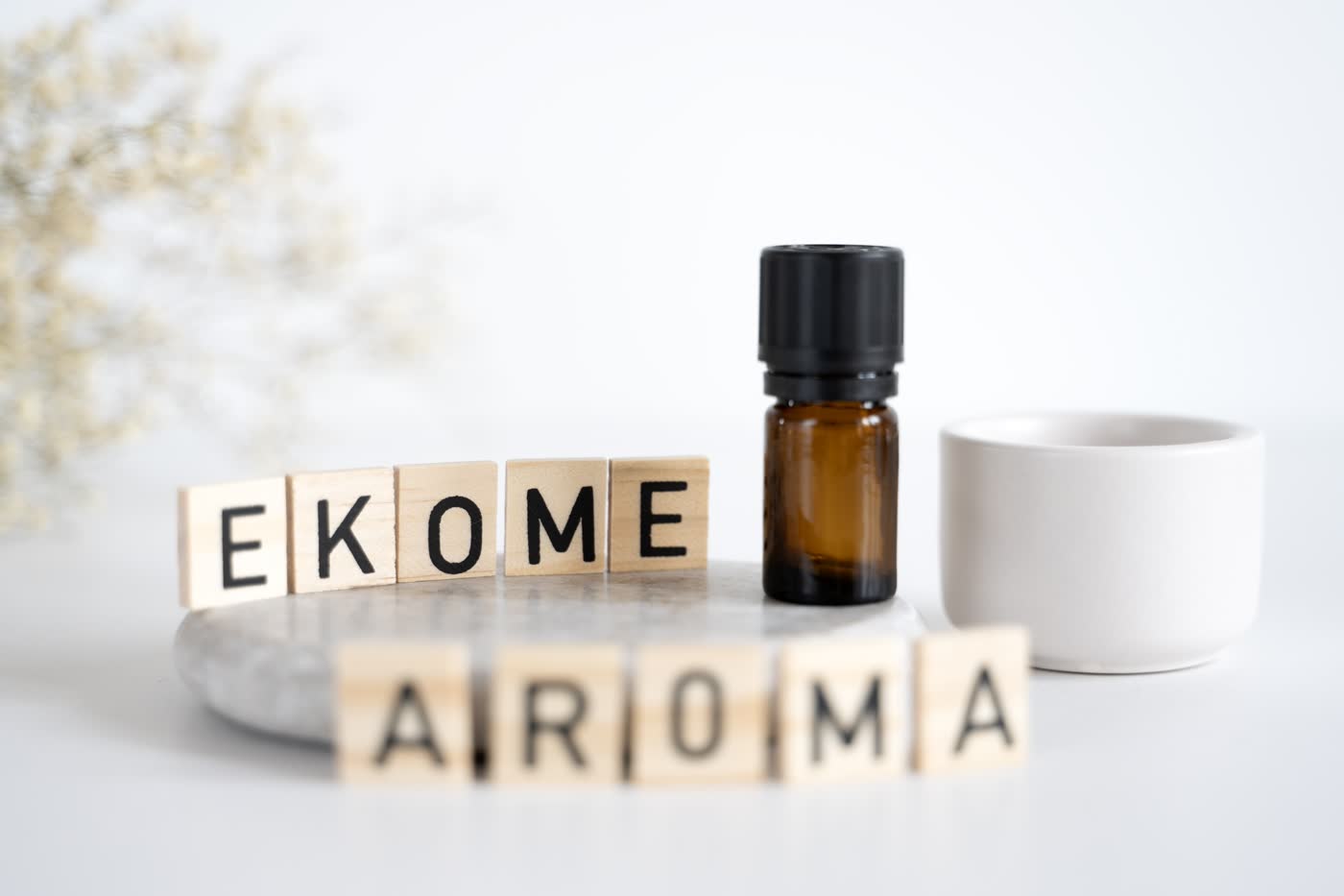 Na pierwszym planie drewniane kostki z literami układającymi się w napisy "EKOME" i "AROMA" na szarym marmurowym tle. W tle niewyraźny, rozmyty bukiet suchych kwiatów, po prawej bursztynowa butelka z czarną nakrętką i po prawej biała ceramiczna miseczka. Całość na jasnym, jednolitym tle.