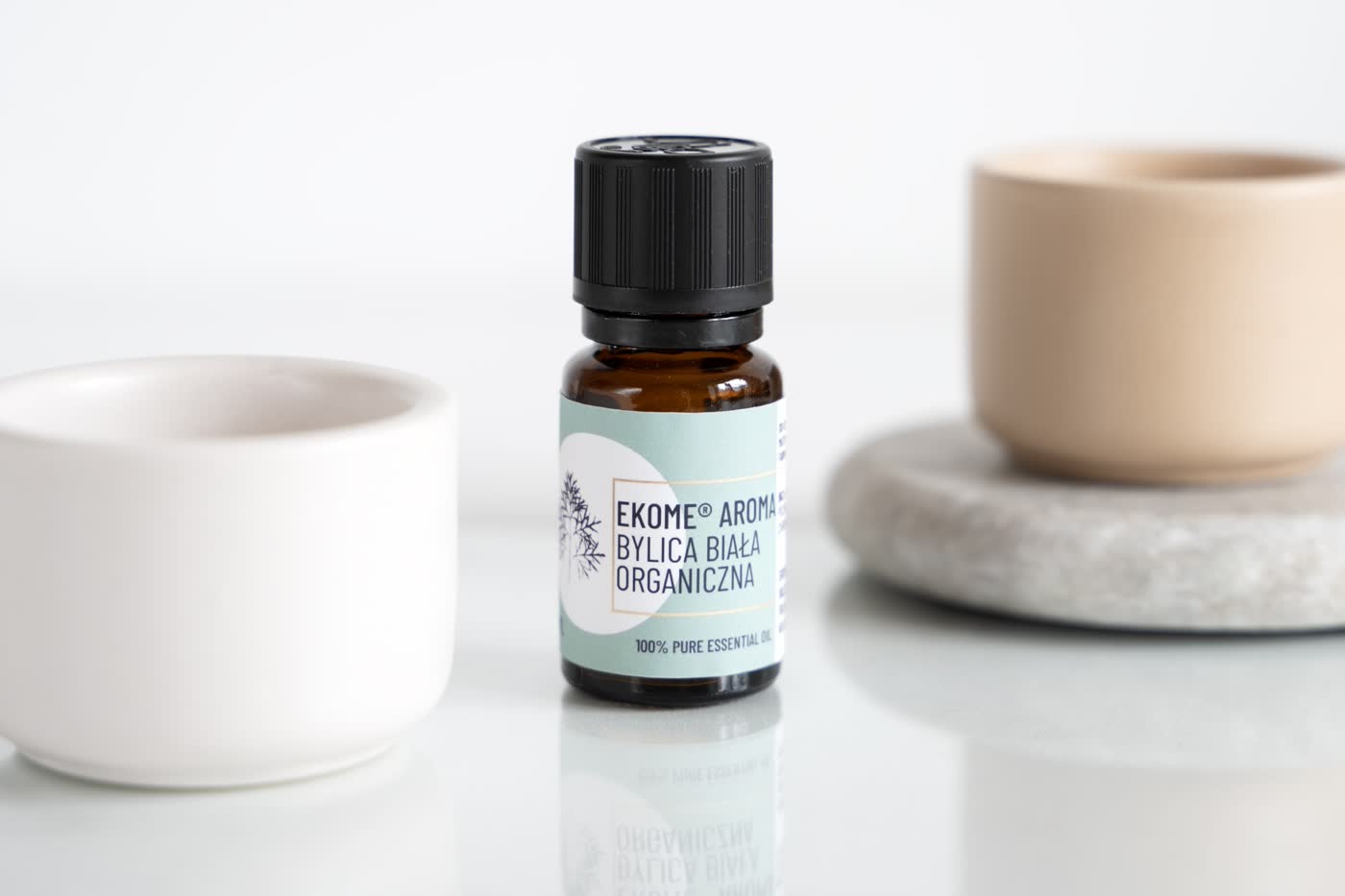 Bursztynowa butelka z olejkiem eterycznym marki EKOME AROMA, z napisem "BYLICA BIAŁA ORGANICZNA" oraz "100% PURE ESSENTIAL OIL". Po lewej stronie znajduje się biała ceramiczna miseczka, a po prawej beżowy kamień i podstawa z szarego kamienia. Tło jest jasne i jednolite, a całość kompozycji utrzymana w delikatnych i naturalnych kolorach.