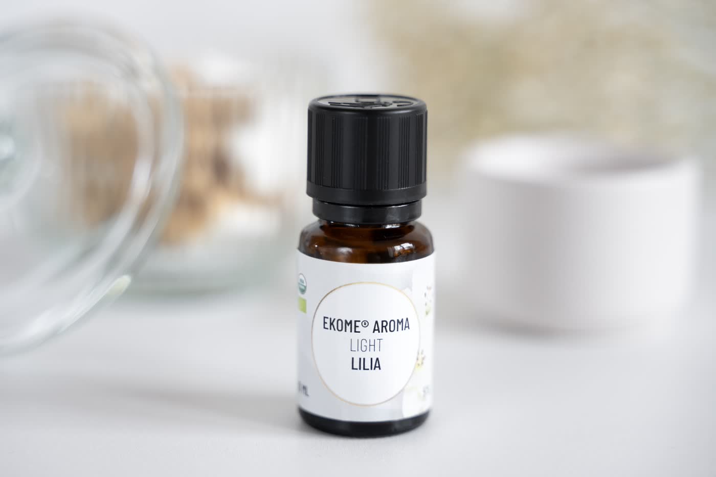 Bursztynowa butelka z olejkiem eterycznym EKOME AROMA "LIGHT LILIA" umieszczona centralnie na zdjęciu. W tle nieostro słoik z korkiem i białe suche kwiaty. Całość na jasnym tle z subtelnie rozmazanymi elementami w tle, co nadaje zdjęciu głębi.