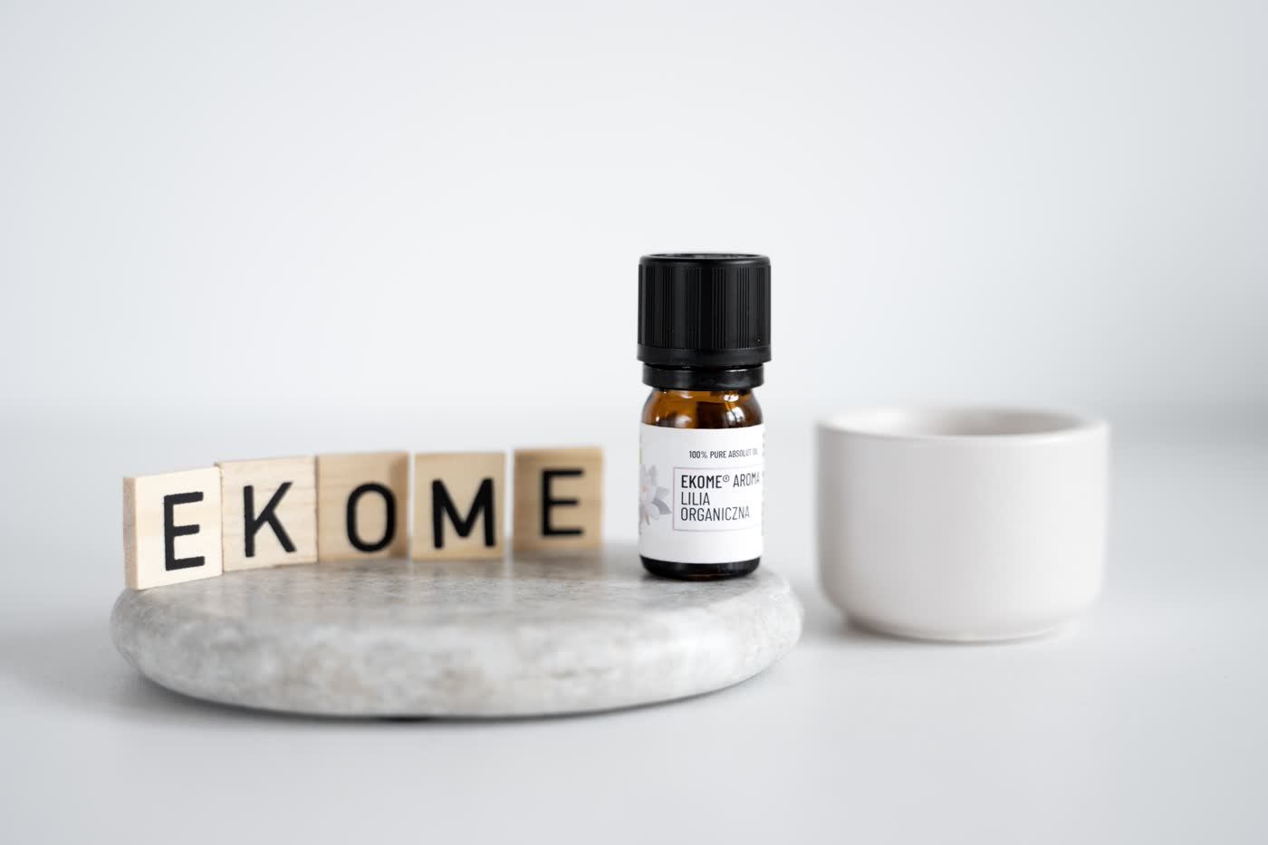 Na pierwszym planie znajduje się bursztynowa butelka z olejkiem eterycznym EKOME AROMA "LILIA ORGANICZNA", umieszczona na szarym kamieniu. Z tyłu ustawione drewniane klocki z literami tworzącymi napis "EKOME". Po prawej stronie biała ceramiczna miseczka. Całość kompozycji na jasnym, jednolitym tle.