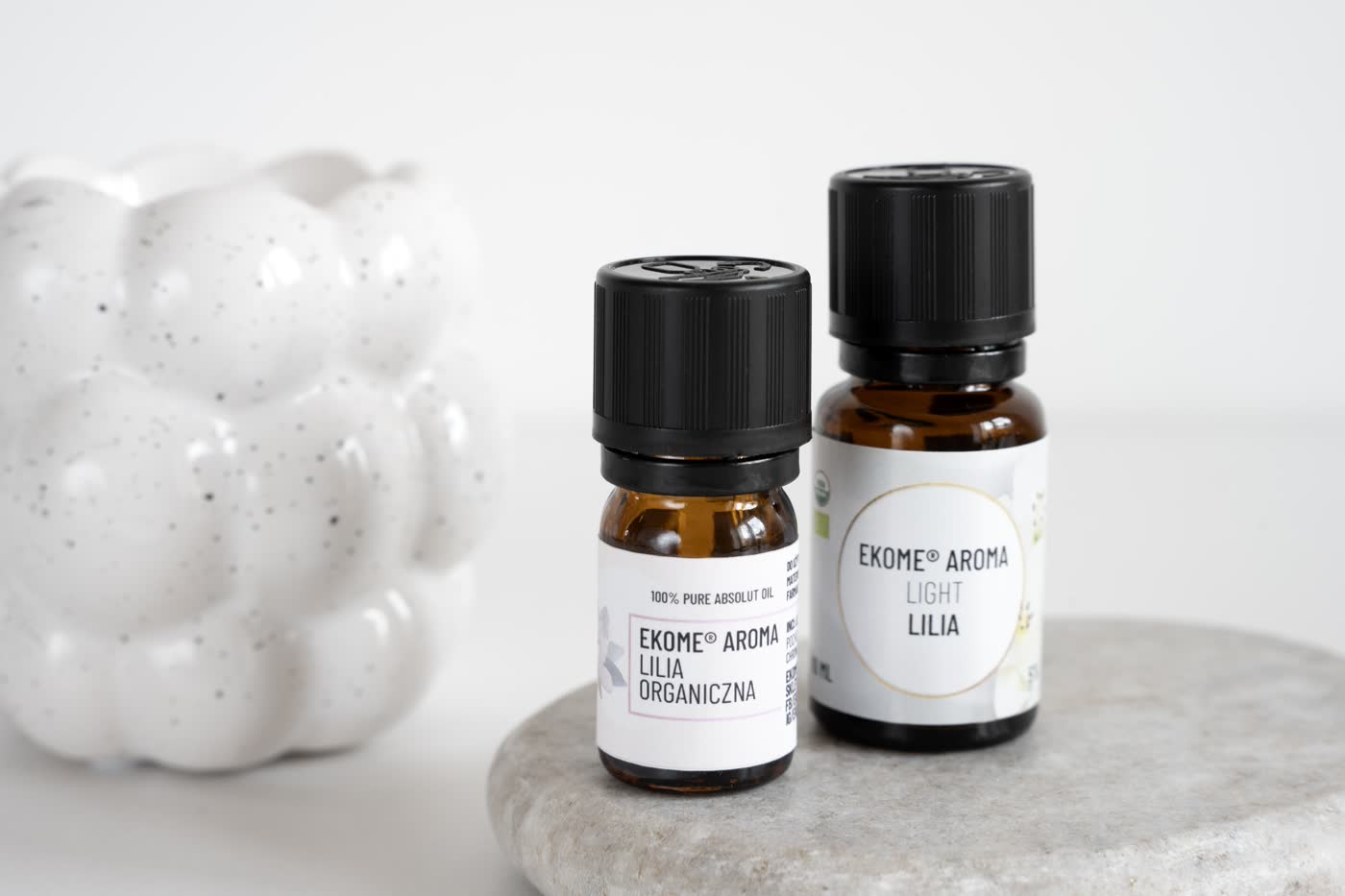 Dwie butelki z olejkami eterycznymi EKOME AROMA na szarym kamieniu. Lewa z etykietą "LILIA ORGANICZNA", prawa "LIGHT LILIA". W lewym górnym rogu nieostre białe ceramiczne figurki. Tło jest jasne i jednolite.