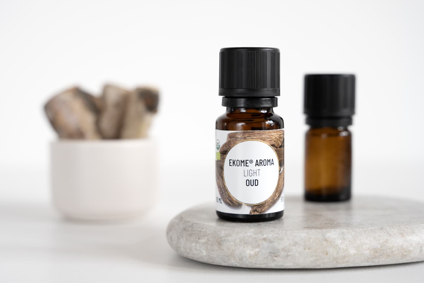 Bursztynowa butelka z olejkiem eterycznym EKOME AROMA "LIGHT OUD" umieszczona na szarym kamieniu. Po prawej stronie druga, rozmyta butelka. W tle biały pojemnik z drewnianymi kawałkami. Całość na jasnym, neutralnym tle z miękkim oświetleniem.
