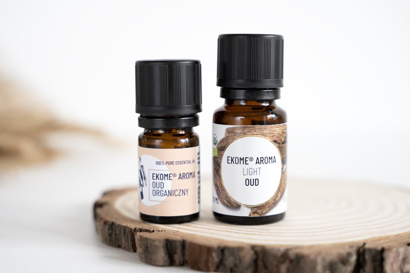 Dwie butelki z olejkami eterycznymi EKOME AROMA umieszczone na przekroju drewnianego pnia. Lewa butelka ma etykietę "OUD ORGANICZNY", prawa "LIGHT OUD". Tło jest jasne i neutralne, a kompozycja podkreśla naturalny charakter produktów.