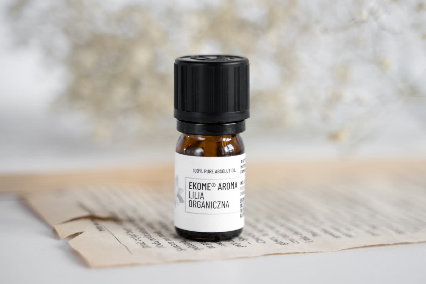Bursztynowa butelka z olejkiem eterycznym EKOME AROMA "LILIA ORGANICZNA" na otwartej książce z widocznym tekstem. W tle rozmyte suche kwiaty. Kompozycja na jasnym tle z naturalnym światłem podkreślającym prostotę i organiczny charakter produktu.
