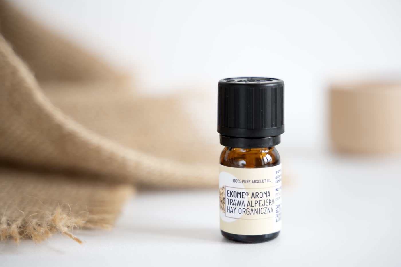 Bursztynowa butelka z olejkiem eterycznym EKOME AROMA "TRAWA ALPEJSKA HAY ORGANICZNA" stoi na białym tle. W tle nieostro jutowy materiał i pastelowy pojemnik. Zdjęcie ma miękkie oświetlenie i naturalne, spokojne kolory.