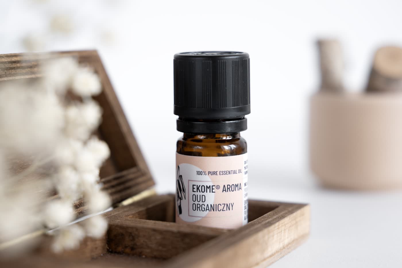Bursztynowa butelka z olejkiem eterycznym EKOME AROMA "OUD ORGANICZNY" na pierwszym planie. Butelka stoi w drewnianej skrzynce, w tle rozmyte białe kwiaty i beżowy ceramiczny pojemnik. Całość na jasnym tle z naturalnymi i neutralnymi barwami.