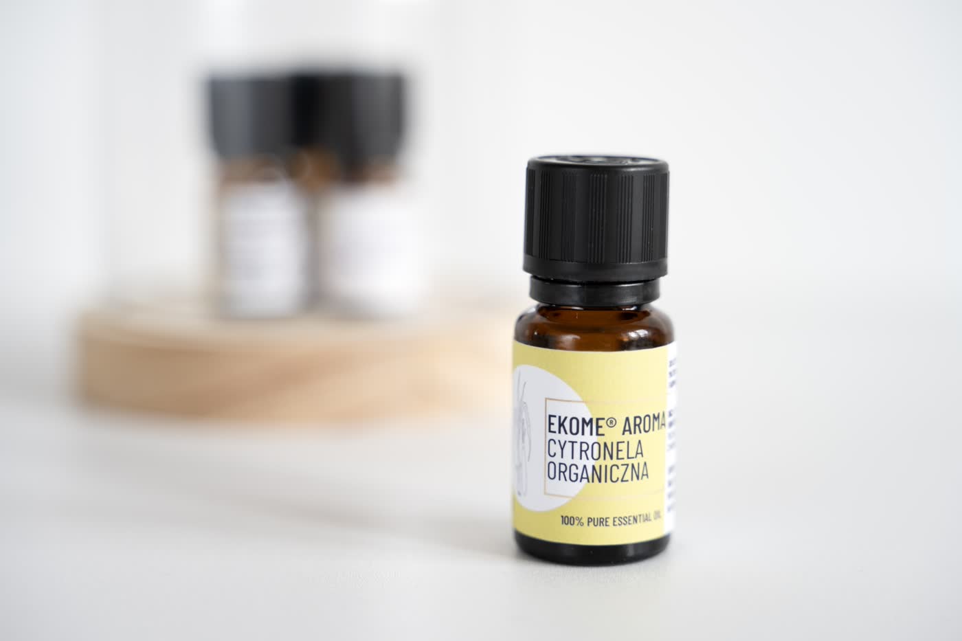 Zdjęcie przedstawia butelkę z olejkiem eterycznym marki "Ekome", z etykietą wskazującą na "Cytronela Organiczna". Produkt jest w centralnym punkcie kadru, z lekko rozmytym tłem, na którym widać inne nieostrzeżone butelki. Butelka olejku jest umieszczona na drewnianej podstawce, a całość ma czyste i minimalistyczne tło, co pozwala skupić uwagę na produkcie. Jasna etykieta z łagodną żółtą barwą sugeruje naturalne pochodzenie i organiczne właściwości olejku.