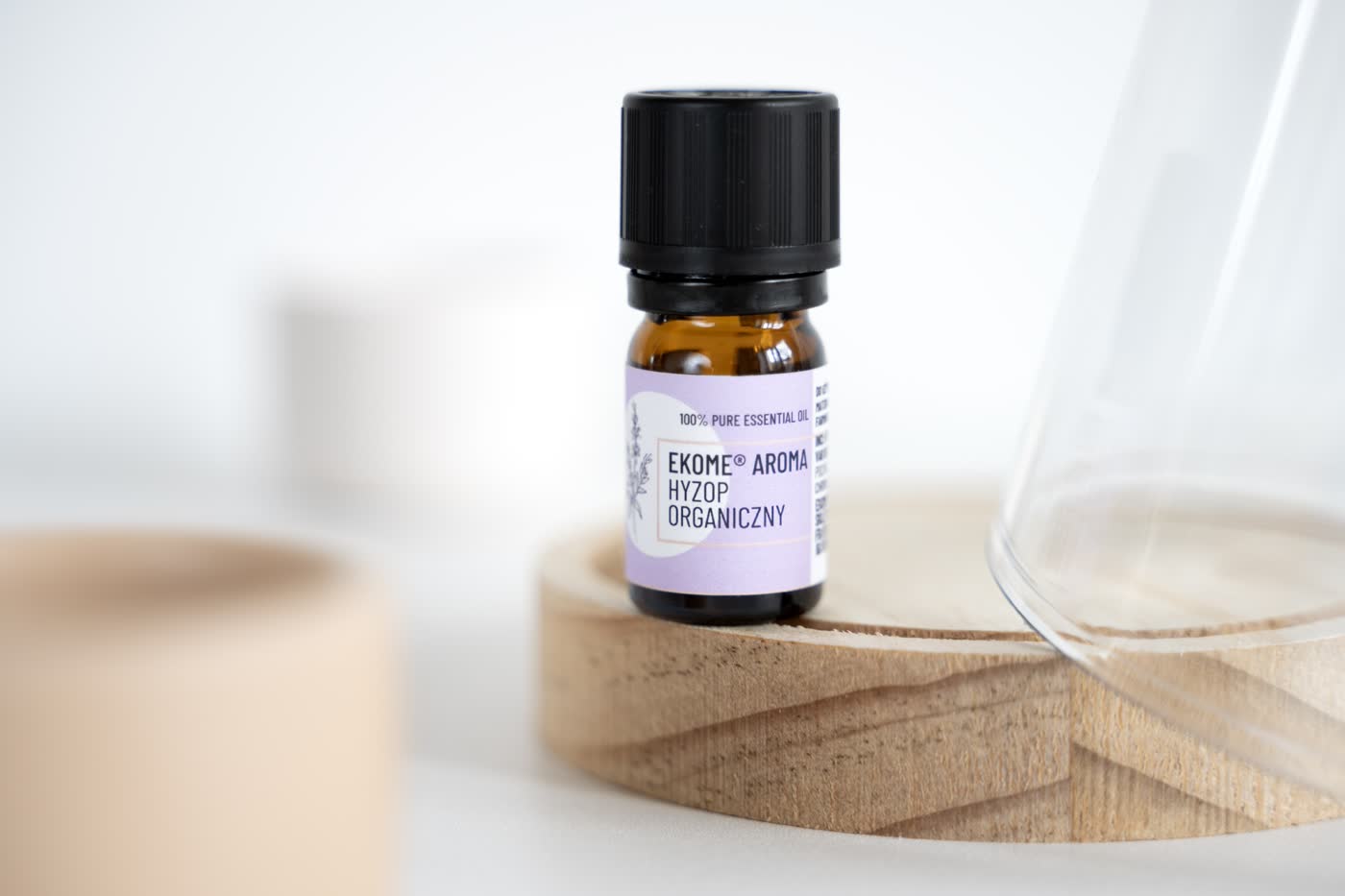 Na pierwszym planie znajduje się butelka z olejkiem eterycznym "EKOME AROMA HYZOP ORGANICZNY" ustawiona na drewnianej podstawce. Tło jest jasne i minimalistyczne, składające się z białych i neutralnych tonów. Z prawej strony zdjęcia pojawia się fragment przezroczystego szklanego naczynia, które dodaje zdjęciu głębi i nowoczesnego charakteru. Na etykiecie butelki widoczne są informacje o produkcie, w tym wyróżnienie 100% czystego olejku eterycznego.