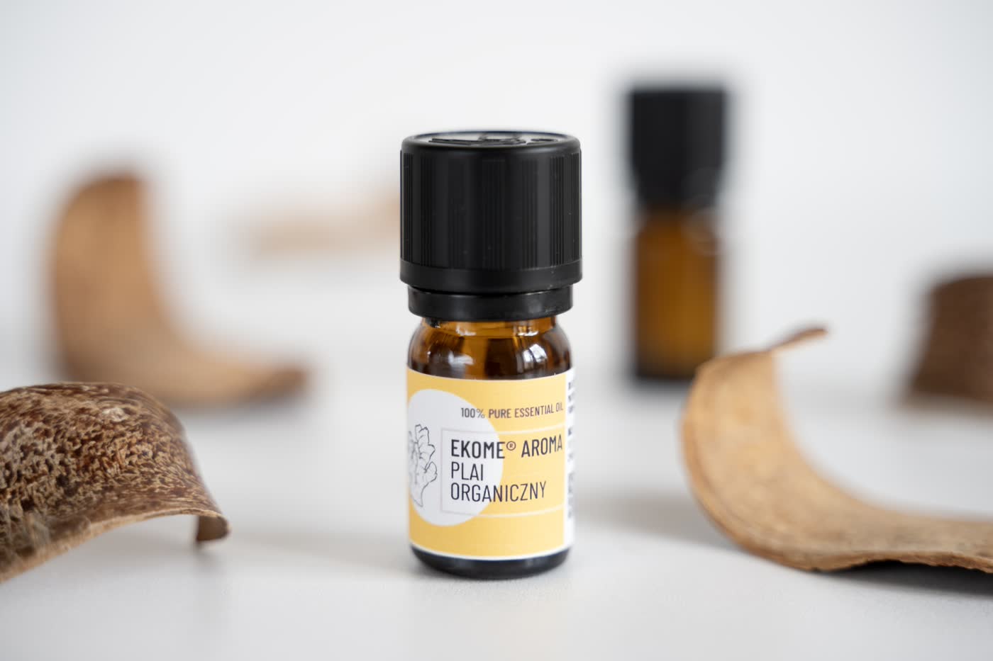 Bursztynowa butelka z olejkiem eterycznym "EKOME AROMA PLAI ORGANICZNY" na pierwszym planie. W tle rozmyte suche łupiny roślinne w ciepłych brązowych tonach na jasnym tle, co nadaje zdjęciu naturalny, ziemisty charakter.