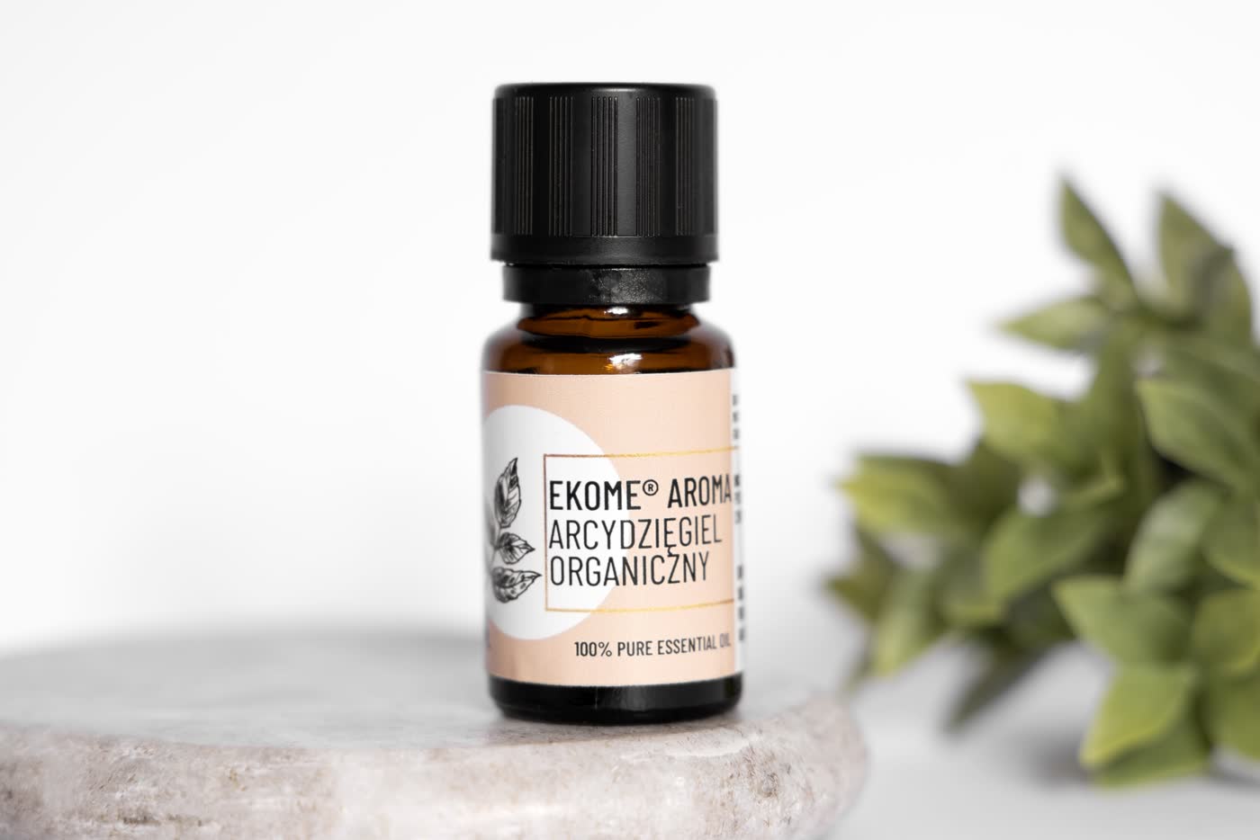 Butelka olejku Ekome Aroma Arcydzięgiel Organiczny na białym podłożu z zielonymi liśćmi w otoczeniu.