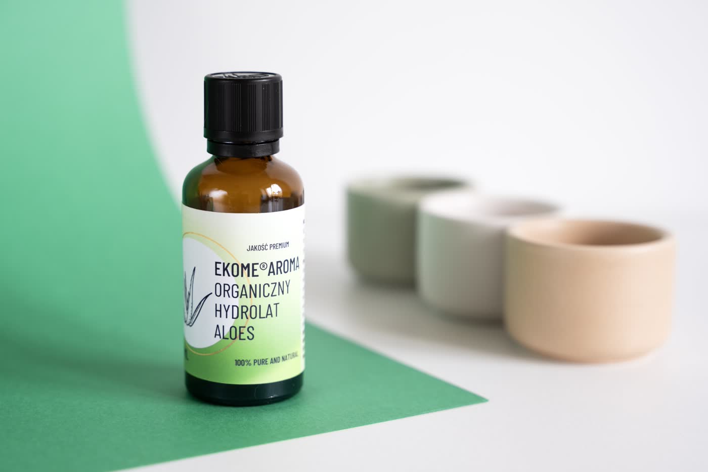 Brązowa butelka z organicznym hydrolatem aloesowym marki Ekome Aroma, oznaczona etykietą z logiem i nazwą, stoi na dwukolorowym tle z odcieniami zieleni i bieli. W tle rozmyte opakowania w ciepłych kolorach beżu i zieleni dodają głębi kompozycji, podkreślając naturalny charakter produktu.