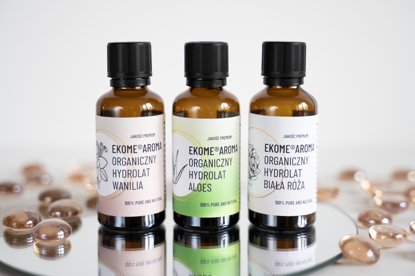Trzy brązowe butelki z organicznymi hydrolatami marki Ekome Aroma. Od lewej: Hydrolat Wanilia, Hydrolat Aloes, Hydrolat Biała Róża. Butelki są umieszczone na lustrzanej powierzchni, co odbija ich obrazy. W tle rozrzucone są przezroczyste kamienie dekoracyjne, dodające elegancji kompozycji.