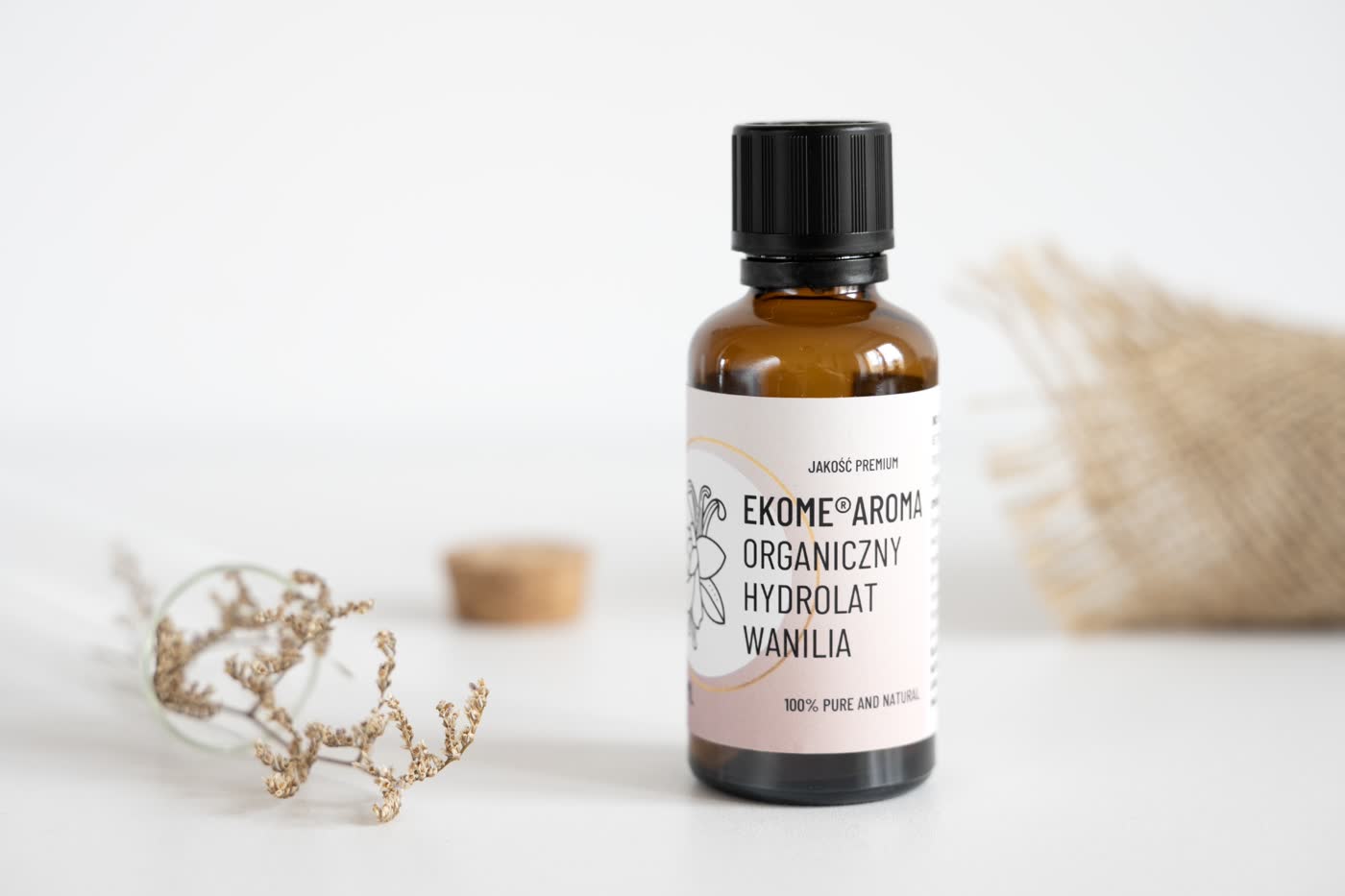 Zdjęcie przedstawia butelkę z brązowego szkła z organicznym hydrolatem o zapachu wanilii marki Ekome Aroma. Etykieta butelki ma kremowe tło z czarnymi i pomarańczowymi akcentami oraz napis "Hydrolat Wanilia". W tle widoczny jest przewrócony szklany słoik z suszonymi kwiatami i jasny materiał.