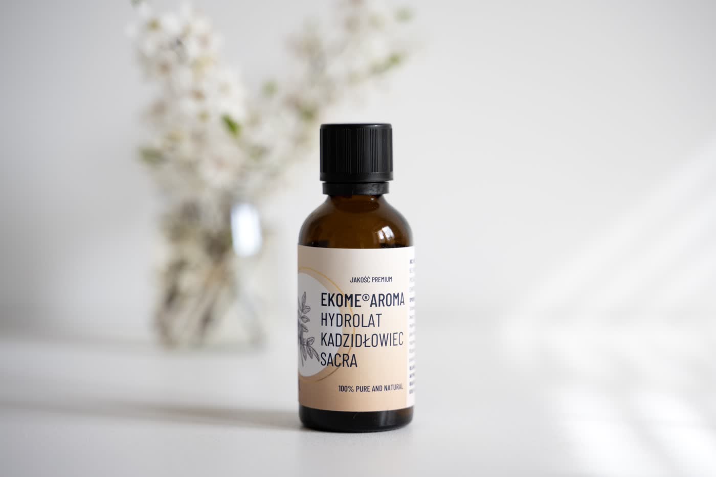 Butelka z brązowego szkła z organicznym hydrolatem kadzidłowiec sacra marki Ekome Aroma stoi na białym tle. Butelka jest zakręcona czarną nakrętką i opatrzona etykietą w odcieniach beżu z nazwą produktu i logo marki. Z tyłu rozmyte kwiaty dodają delikatności i estetycznego wyglądu kompozycji. Całość prezentuje czystą i spokojną atmosferę, akcentując naturalność produktu.