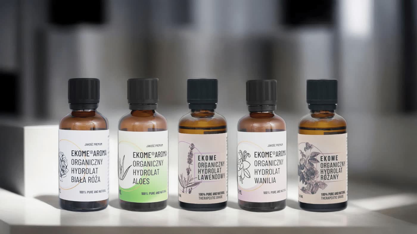 Na zdjęciu znajduje się seria brązowych butelek z hydrolatami organicznymi marki Ekome Aroma. Z lewej strony widać "Organiczny Hydrolat Biała Róża", obok "Organiczny Hydrolat Aloes", w środku "Organiczny Hydrolat Lawendowy", następnie "Organiczny Hydrolat Wanilia" i na końcu "Organiczny Hydrolat Różany". Buteleki są ustawione na białym tle z rozmytymi kształtami w tle, co skupia uwagę na produktach.