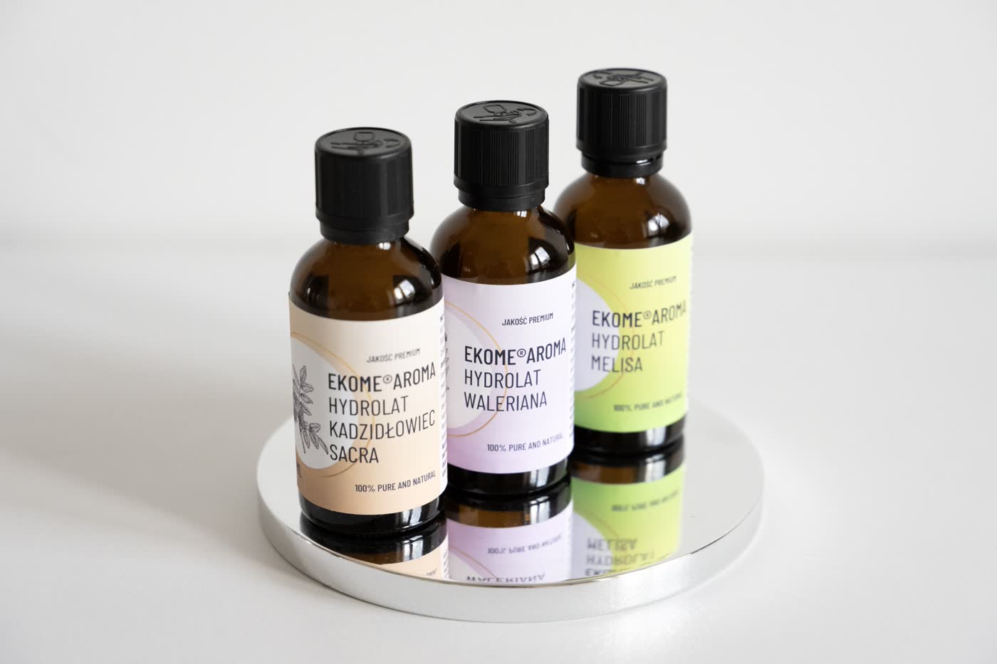 Trzy butelki organicznych hydrolatów marki Ekome Aroma stoją na okrągłym, lustrzanym podkładzie, który odbija ich obrazy. Od lewej znajduje się hydrolat kadzidłowiec, w środku waleriana, a po prawej melisa. Butelki mają czarne nakrętki i etykiety z nazwą oraz grafiką roślin, z których zostały wyprodukowane. Całość prezentuje się na białym tle, co podkreśla naturalne barwy etykiet i eleganckie opakowanie produktu.