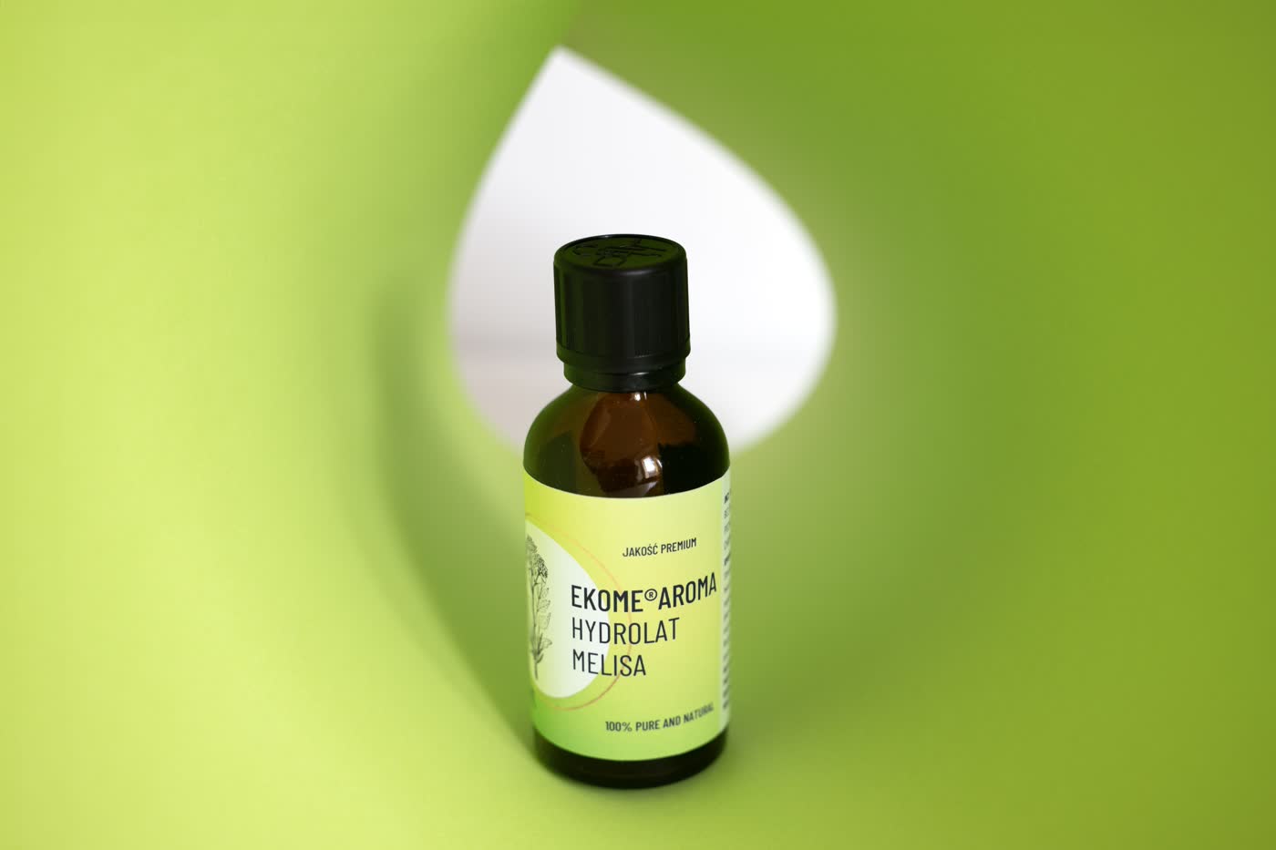 Butelka z ciemnego szkła z organicznym hydrolatem melisy marki Ekome Aroma, z czarną nakrętką i etykietą w pastelowych zielonych odcieniach, stoi na jednolitym, intensywnie zielonym tle z białym akcentem w kształcie łezki, co dodaje zdjęciu nowoczesnego charakteru.