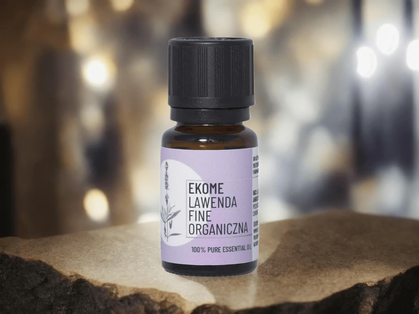Mała butelka olejku "EKOME LAWENDA FINE ORGANICZNA" na kamiennym blacie. Brązowa butelka z etykietą fioletowo-białą stoi na naturalnym kamieniu. W tle nieostra złota poświata daje ciepły klimat.