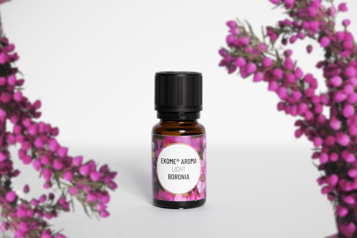 Ekome® Aroma Light Boronia – buteleczka olejku absolutnego z boronii na jasnym tle, otoczona rozmytymi, fioletowymi kwiatami