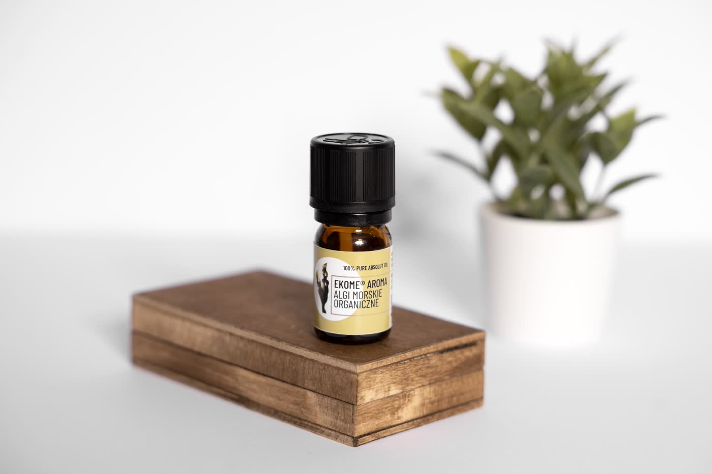 Butelka olejku Ekome Aroma Algi Morskie Organiczne ustawiona na drewnianym pudełku, w tle zielona roślina w doniczce.
