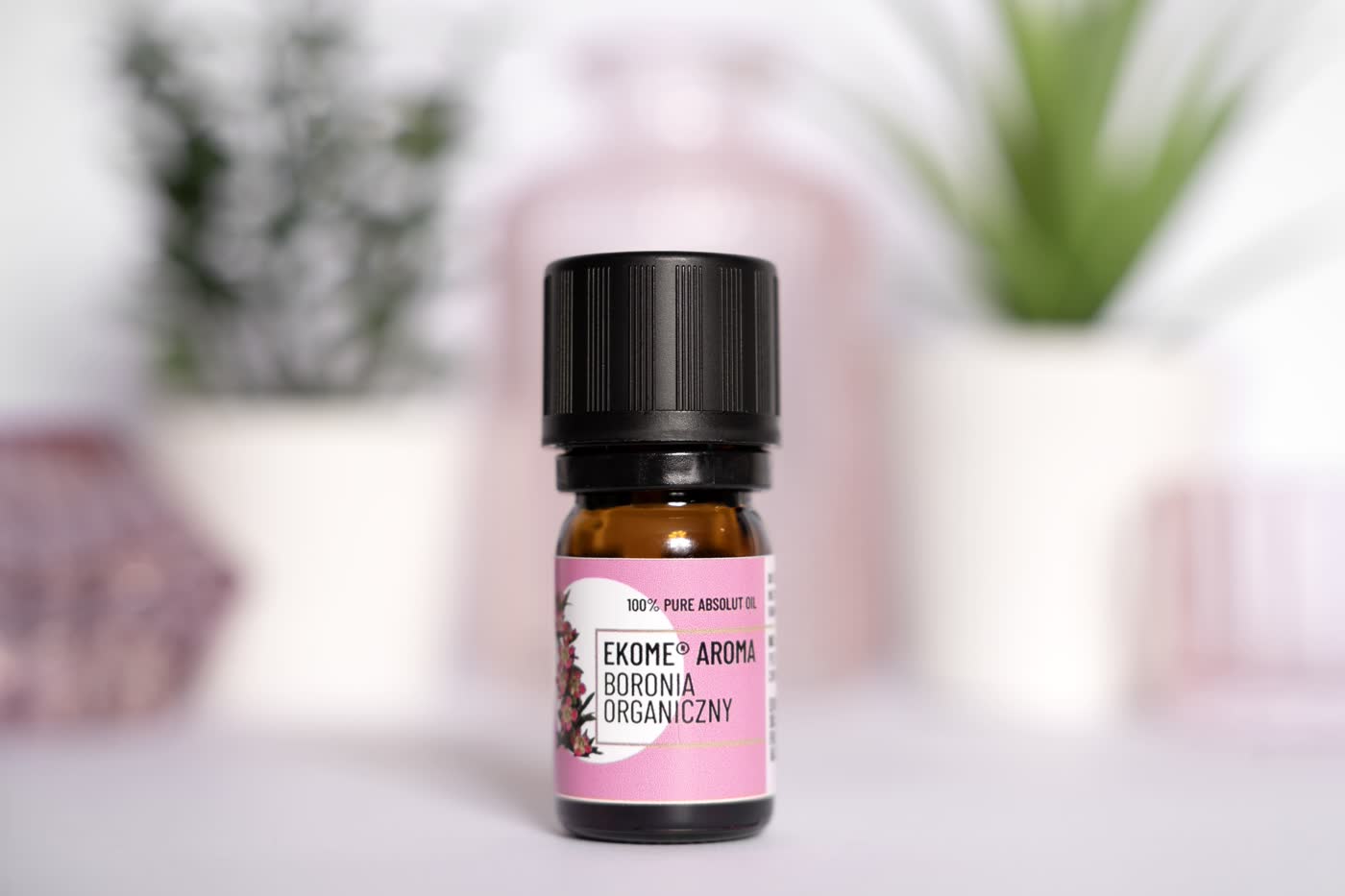 Ekome® Aroma Boronia Organiczny, 100% olejek absolutny, w szklanej buteleczce, ustawiony na jasnym tle z rozmytym tłem roślinnym w tle.