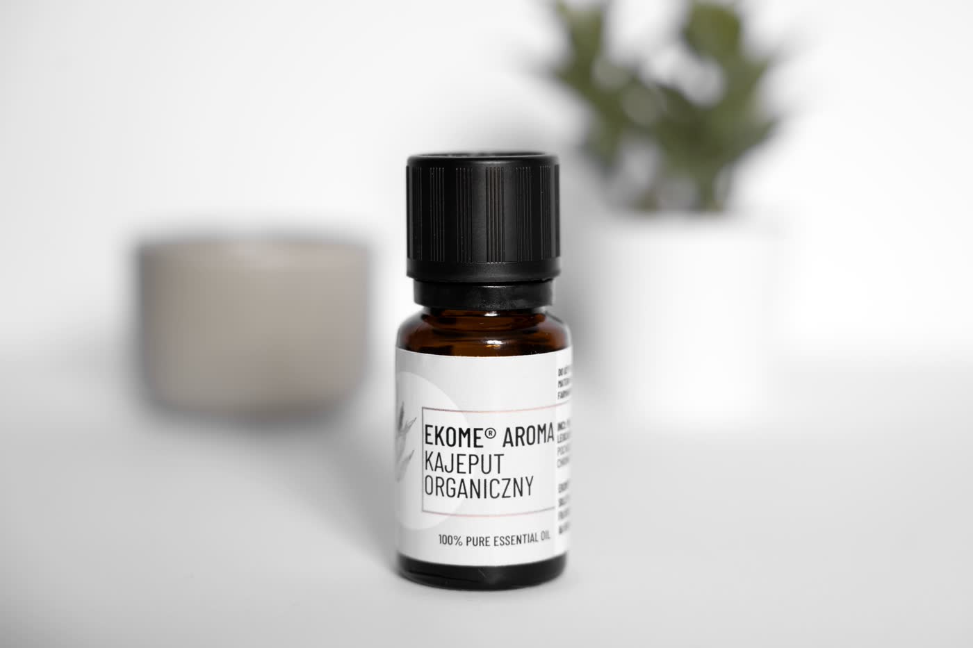 Butelka olejku Ekome Aroma Kajeput Organiczny na jasnym tle z subtelnymi elementami dekoracyjnymi w tle.