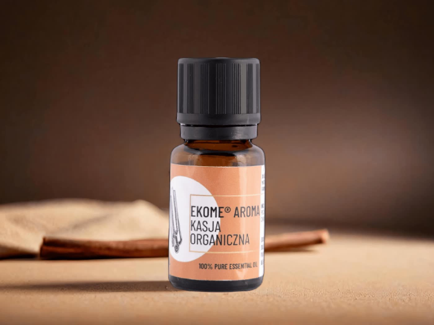 Butelka Ekome® organiczny olejek eteryczny Kasja 10 ml na tle drewnianego stołu, z korą kasji w tle. Produkt w szklanej butelce z czarną nakrętką i brązową etykietą.