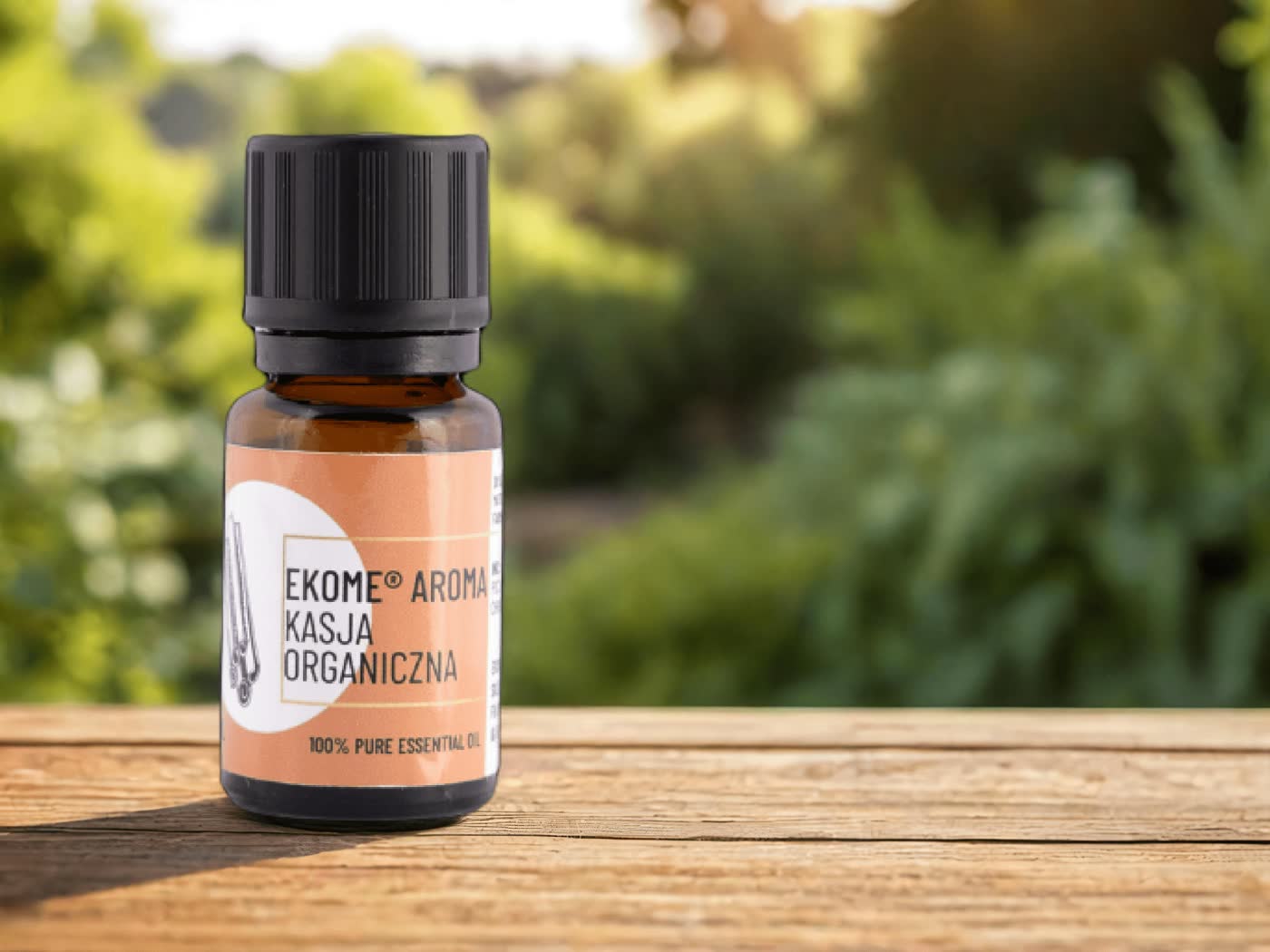 Butelka Ekome® organiczny olejek eteryczny Kasja 10 ml na tle bujnej zieleni ogrodu, na drewnianym stole. Produkt w szklanej butelce z etykietą w odcieniach brązu, czarną nakrętką.
