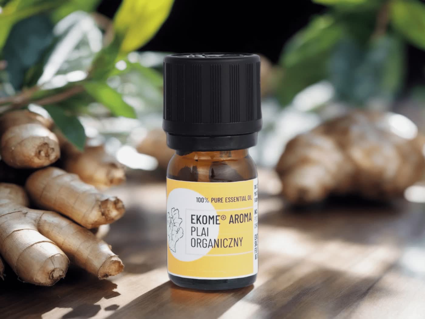 Bursztynowa butelka z olejkiem eterycznym "EKOME AROMA PLAI ORGANICZNY" stoi na drewnianym stole. W tle naturalne korzenie imbiru i zielone liście, które dodają zdjęciu świeżości i podkreślają organiczny charakter produktu.