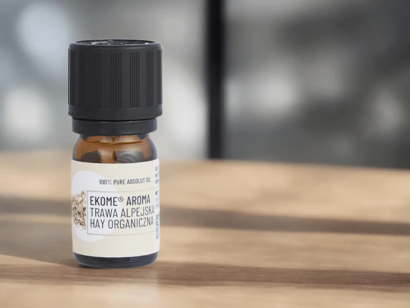 Bursztynowa butelka z olejkiem eterycznym oznaczona jako "EKOME AROMA TRAWA ALPEJSKA HAY ORGANICZNA" stoi na drewnianym blacie. Tło jest rozmyte, z delikatnymi odcieniami szarości, co pozwala skupić uwagę na butelce.