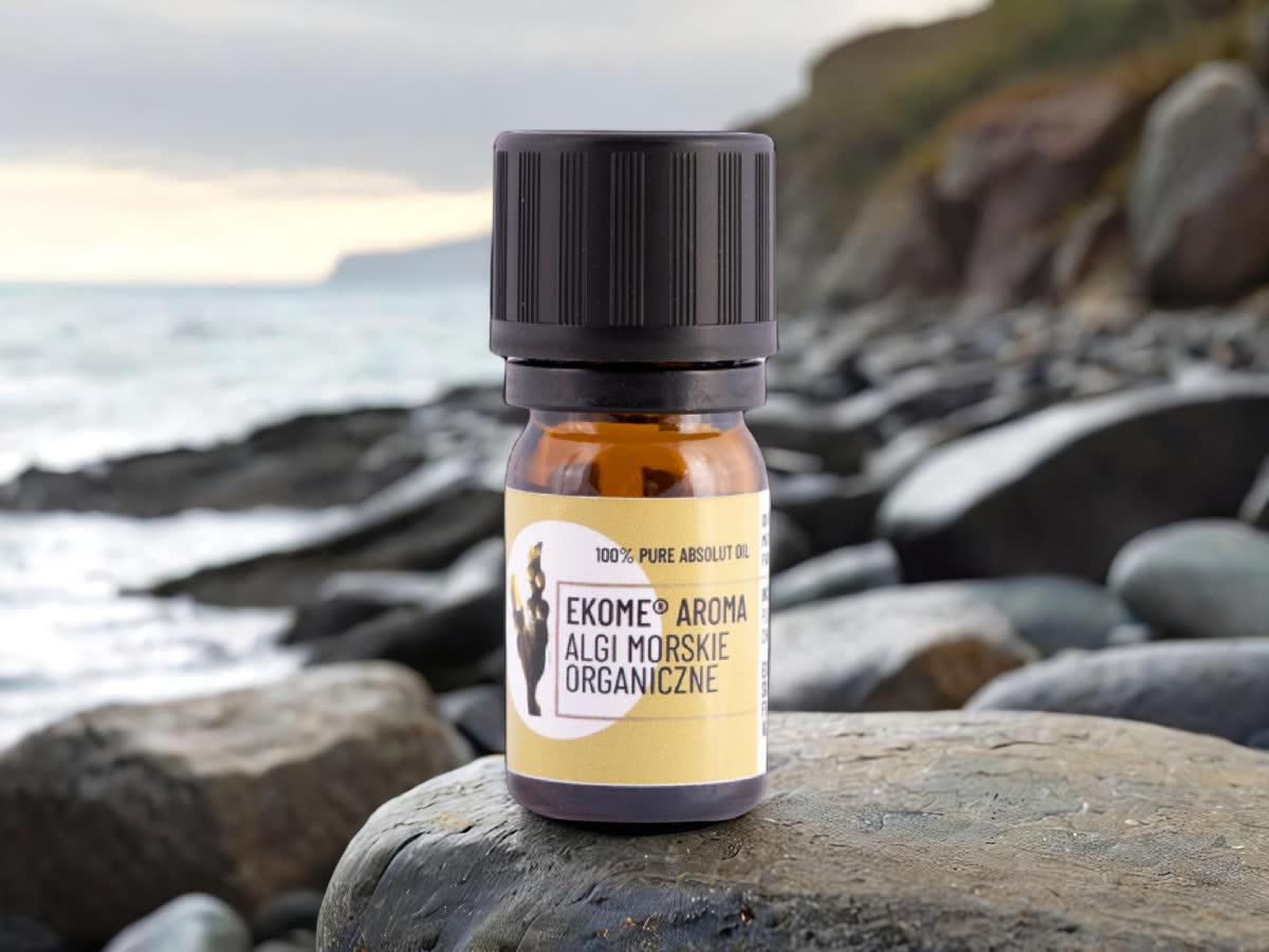 Szklana butelka z olejkiem absolutnym „Ekome® Aroma Algi Morskie Organiczne” ustawiona na skalistym brzegu morza, na tle fal i klifów w naturalnym, przybrzeżnym krajobrazie.
